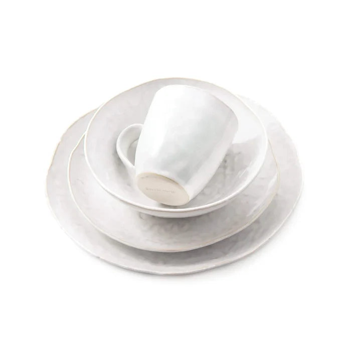 A neatly stacked set of Simon Pearce’s Burlington Dinnerware Cloud is displayed on a white background, including a mug, bowl, salad plate, and an organically edged dinner plate. Each white ceramic piece features tactile uniqueness with subtle freehand pinch-pot design details.