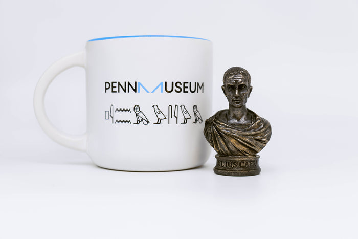 Caesar bust displayed next to white mug with Penn museum logo and hieroglyphs underneath logo.