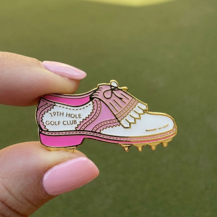 A hand with pink manicured nails holds a small enamel pin shaped like a pink and white golf shoe with gold metal outlines. The pin reads '19TH HOLE GOLF CLUB' and is photographed against an out-of-focus green background.