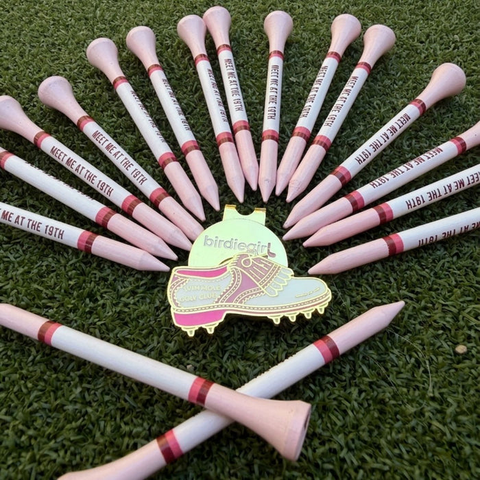 A golf-themed flat lay featuring a central gold-tone shoe-shaped charm engraved with 'birdiegirl' and pink-and-white tees arranged in a circle on artificial green grass. Several tees are tipped with round pink golf tees, and one tee at the bottom is tipped with a pink tee cone.