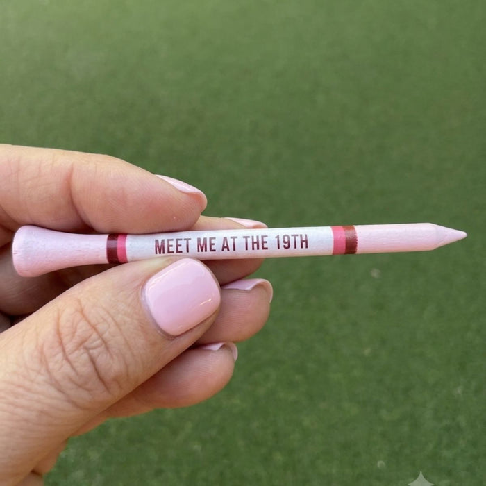 A hand holding a pink golf tees marker that reads 'MEET ME AT THE 19TH' against a green grass background.
